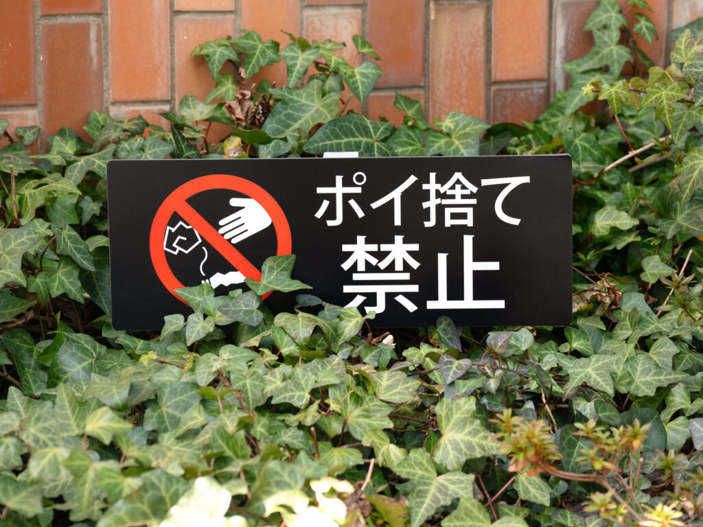 A black sign reading ポイ捨て禁止 (No littering) set among green ivy in front of a brick wall.