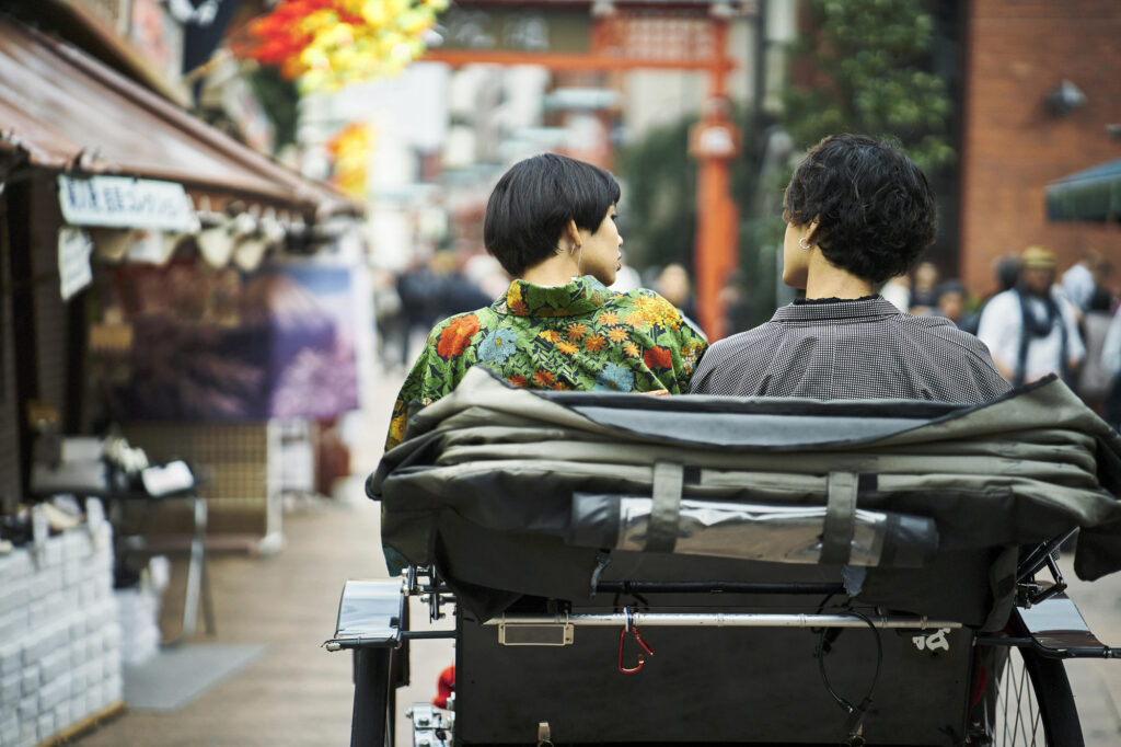 Two passengers, one in a floral kimono, ride a rickshaw down a shopping street toward a red torii gate
