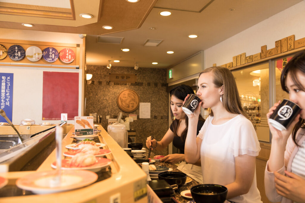 Three diners drink green tea at a conveyor-belt sushi counter, with plates and price placards nearby