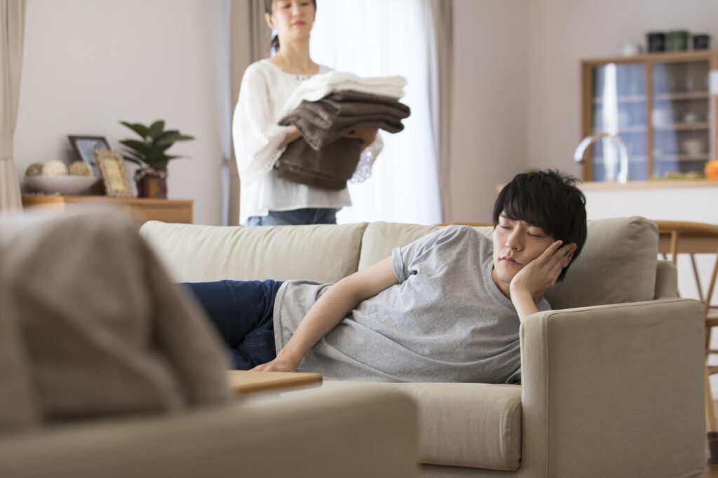 A man reclines on a sofa with his eyes closed while a woman stands behind him holding folded laundry.