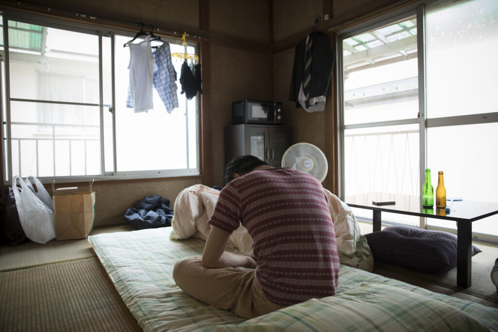 A man sits hunched over on a futon in a cluttered room with laundry hanging by the window