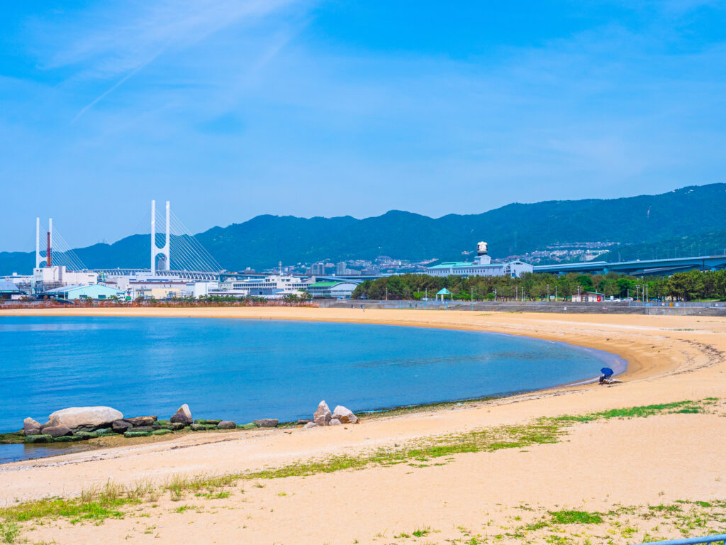 The curved man-made shoreline of Shio-Ashiya Beach, with calm bay water and mountains on the horizon.