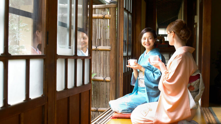 Two women in kimono sit on a traditional wooden veranda holding cups of tea and smiling in conversation
