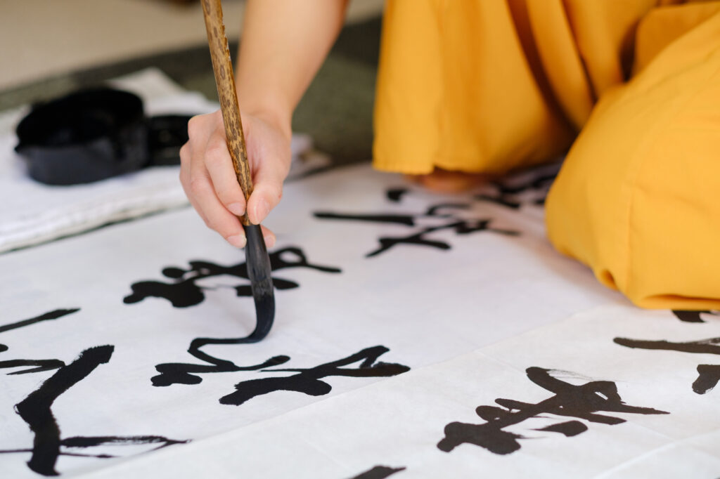 A person in an orange kimono writes large kanji on paper with an ink brush
