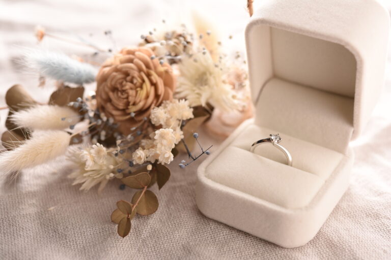 A diamond solitaire ring in an open ivory box beside a bouquet of dried flowers