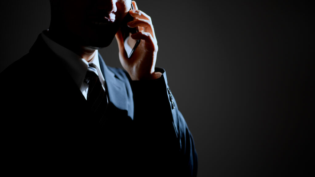 A suited man stands in shadow, holding a smartphone to his ear against a dark background
