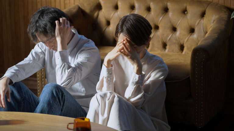 Distressed couple sitting on the floor against a leather sofa, each holding their head in their hands