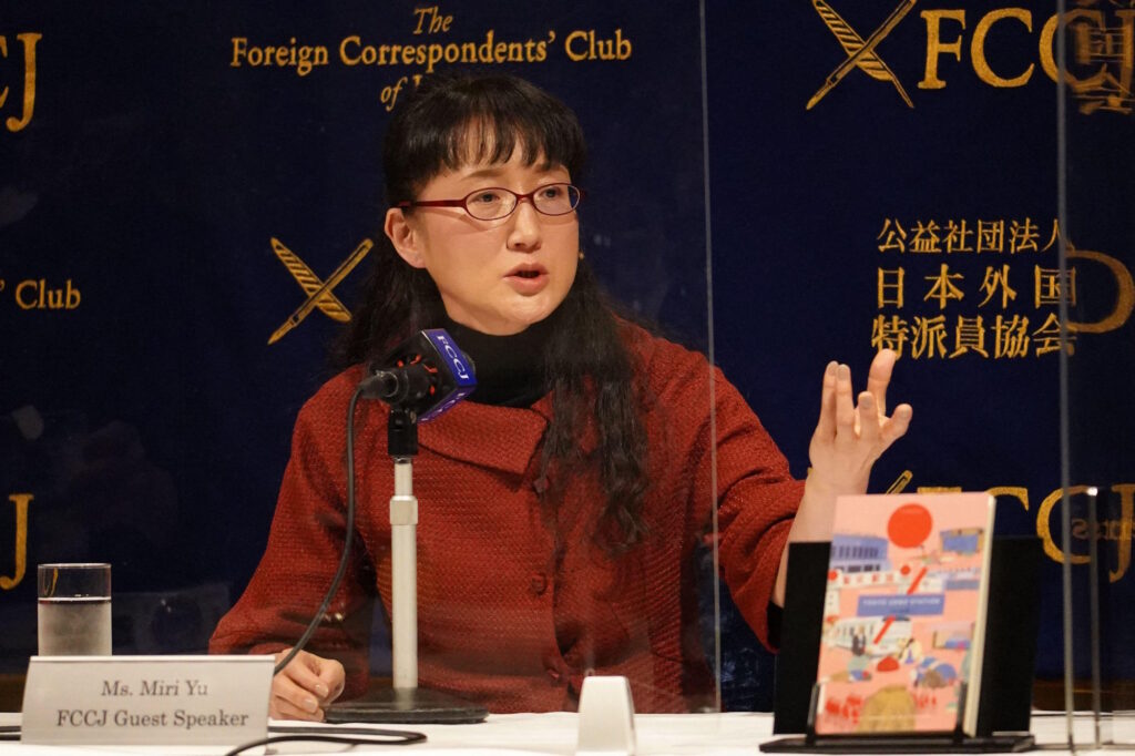 Yu Miri speaking at a microphone at the Foreign Correspondents Club of Japan, wearing a red jacket