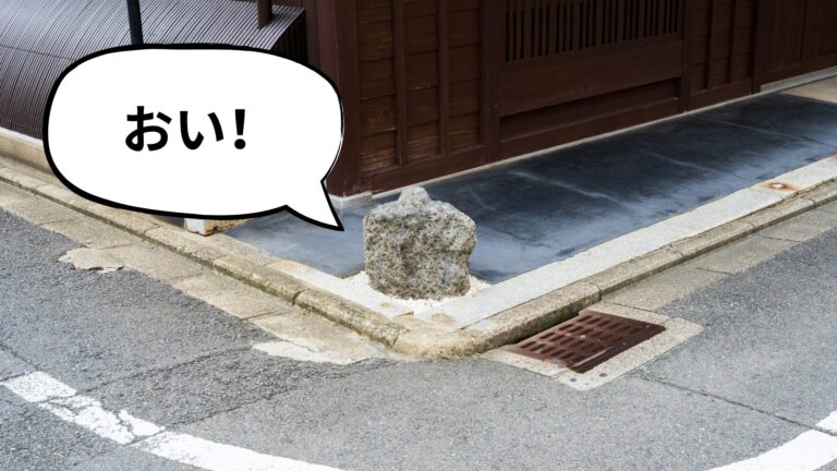 A rough granite ikezu-ishi stone at a wooden Kyoto machiya's corner, with a cartoon speech bubble reading "おい!" ("Hey!")
