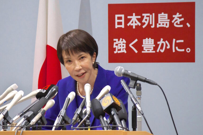 Takaichi Sanae speaking behind a bank of microphones, a Japanese flag and red LDP slogan sign behind her