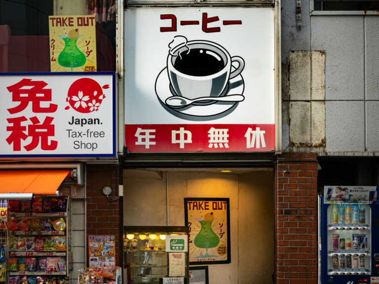 Coffee Shop Cool's entrance sign, an illustrated coffee cup, above its Kabukicho doorway next to a tax-free drugstore.