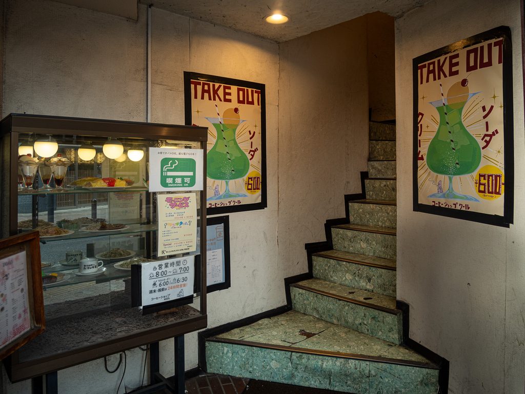 Stairwell up to Coffee Shop Cool, with a lit food-sample case, cream soda takeout posters, and a smoking-permitted sign.