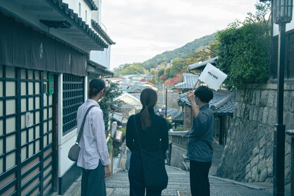 Two guests follow a guide holding a MUJI BASE flag down a stone-stepped Kyoto lane