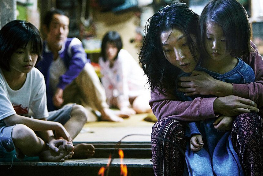 A woman holds a young girl close as the makeshift family sits together in their cramped home, a still from the film Shoplifters.