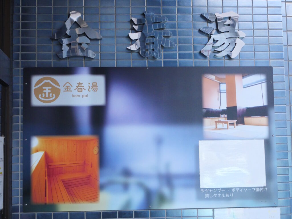 Polished metal 金春湯 sign above kom-pal blue-tiled entrance, beside a board showing its sauna and tatami lounge