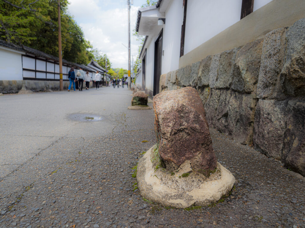 A weathered ikezu-ishi stone set in concrete at the foot of a walled Kyoto lane, with pedestrians walking in the distance