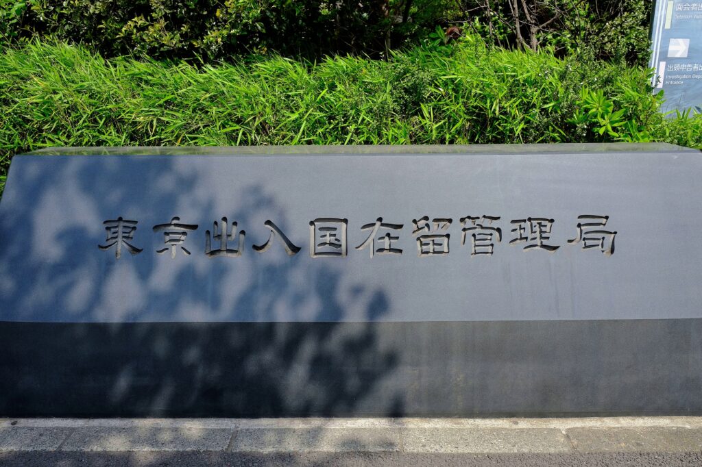A dark stone sign engraved '東京出入国在留管理局' (Tokyo Regional Immigration Bureau), backed by green foliage.
