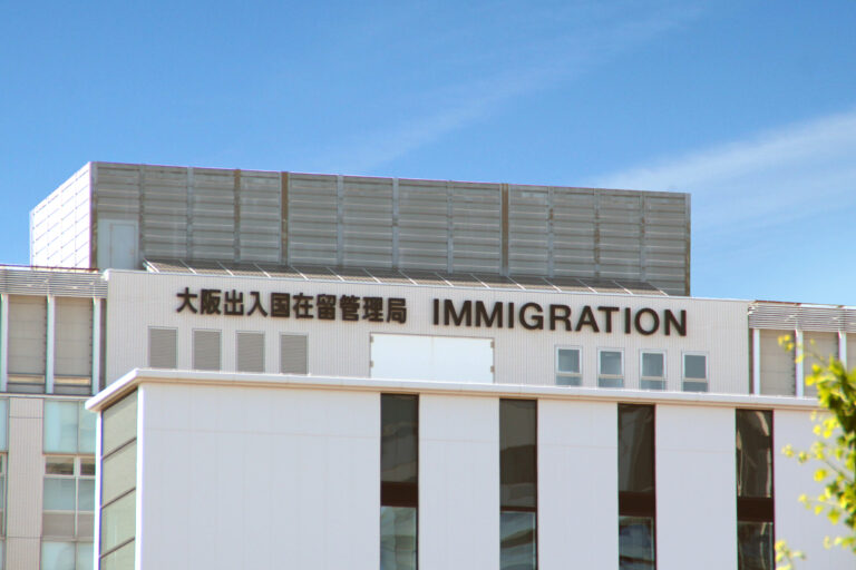 The Osaka Regional Immigration Bureau, its rooftop sign reading '大阪出入国在留管理局 IMMIGRATION,' under a blue sky.