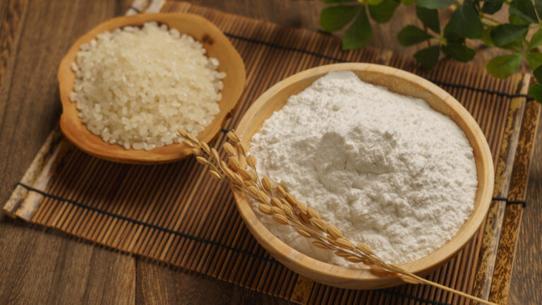 Bowls of uncooked white rice and white rice flour with rice stalks, on a bamboo mat.