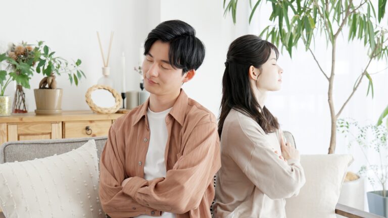 A young couple sit back-to-back on a sofa with arms crossed and eyes closed in a bright living room