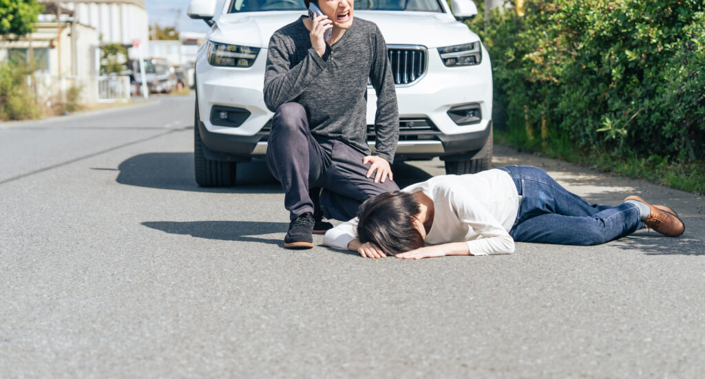 A man kneeling on a road phones for help beside a person lying in front of a white car
