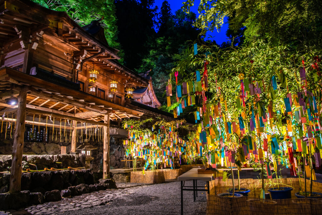 Kifune Shrine at dusk, its wooden hall lit by lanterns beside bamboo hung with colorful paper strips
