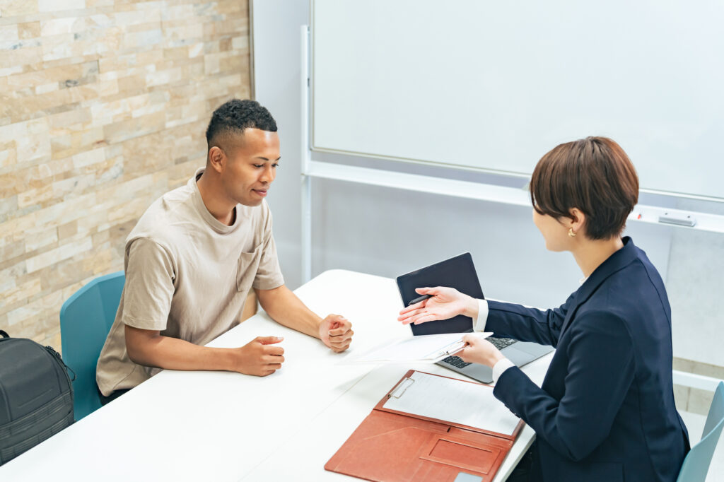 A man talks with a suited advisor holding documents and a laptop across an office desk