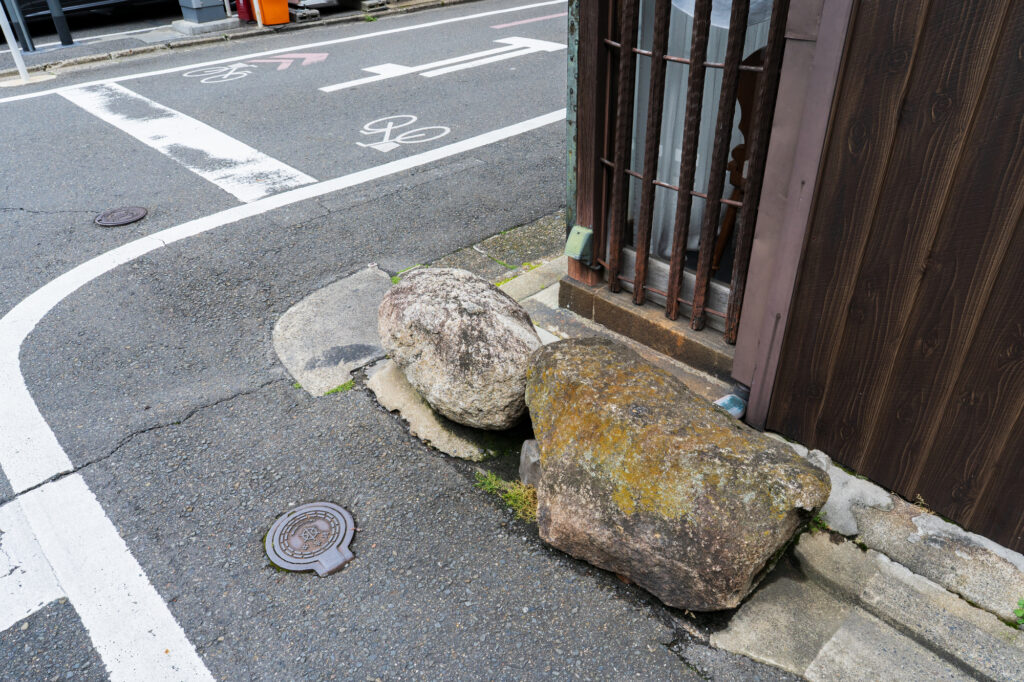 Two large weathered ikezu-ishi stones set at the corner of a wooden building beside a narrow Japanese street.