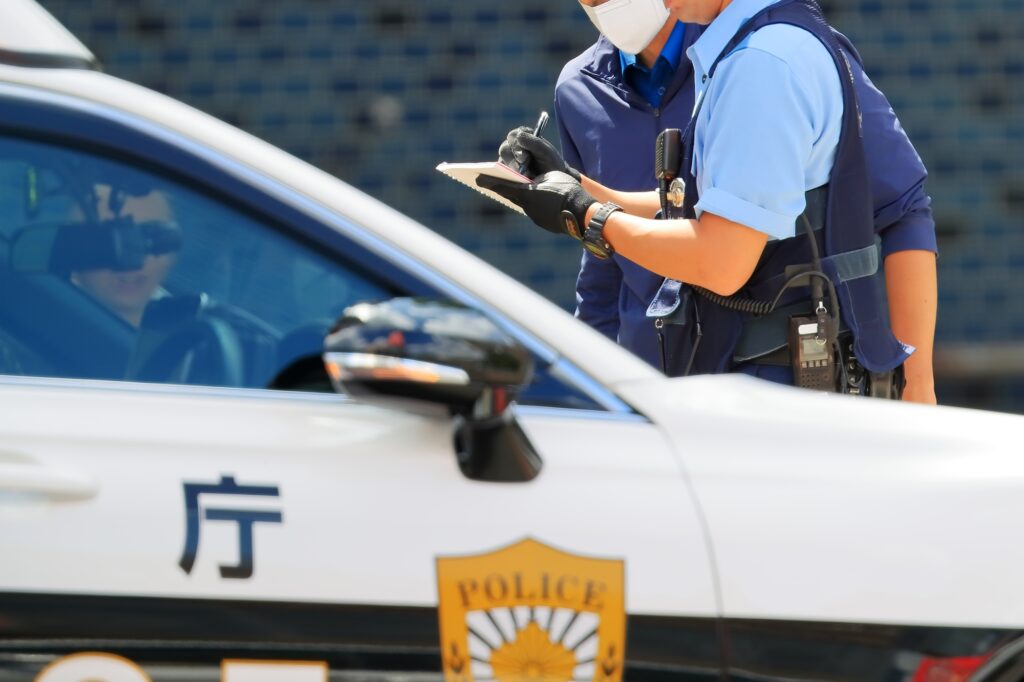 A Japanese police officer writing in a notebook beside a white patrol car marked POLICE