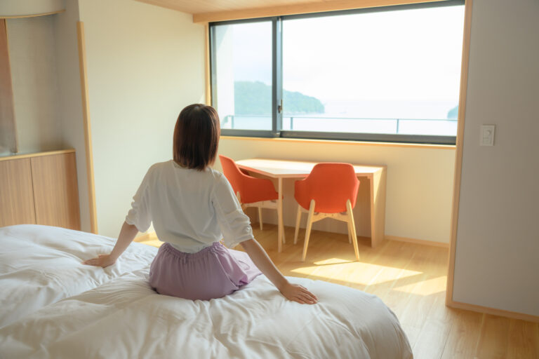 Woman on a hotel bed looking out a wide window at a coastal sea view, beside a wooden table and two orange chairs