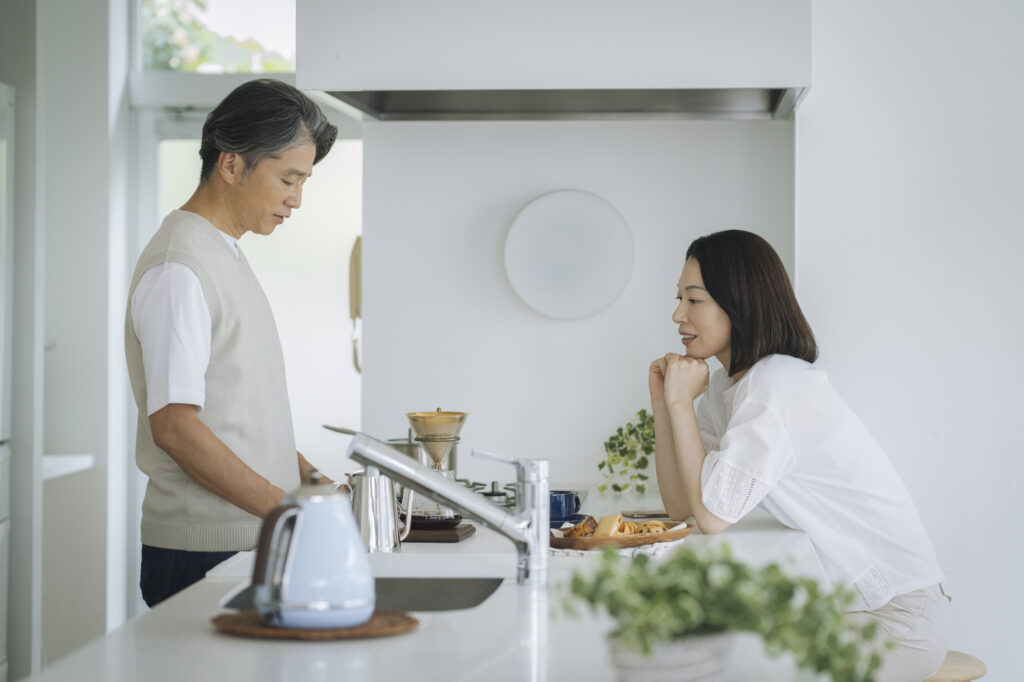 A middle-aged couple talk in a bright kitchen as the man prepares pour-over coffee at the counter