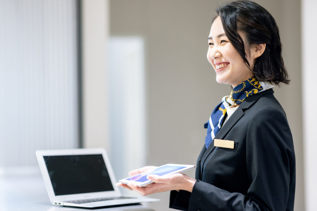 Smiling hotel receptionist in a dark uniform holding a tablet at a front desk with a laptop
