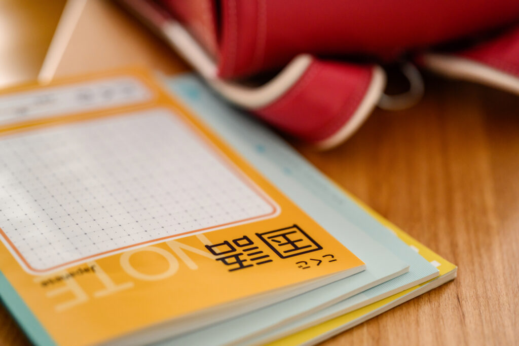 A Japanese kokugo (national language) exercise notebook with grid squares, resting on a wooden floor beside a red bag.