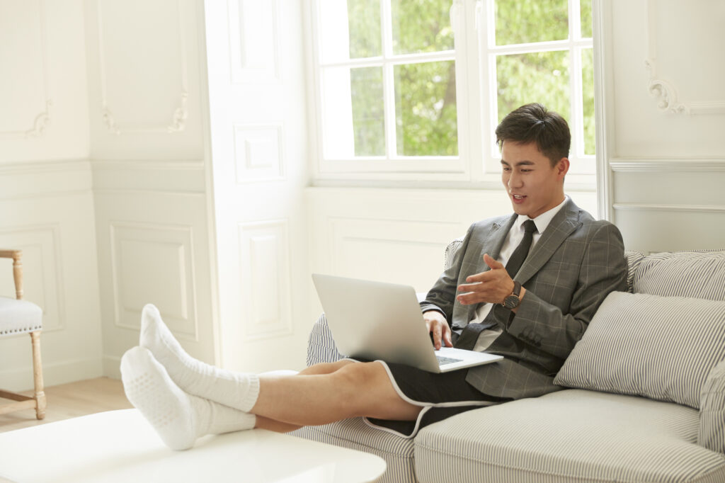 A man in a suit jacket and tie on top but shorts and socks below works on a laptop with his feet up.