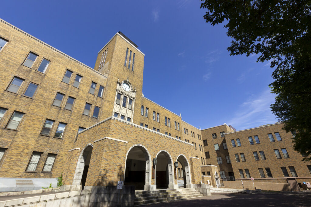 A historic yellow-brick building with a clock tower and arched arcade entrance on the Hokkaido University campus