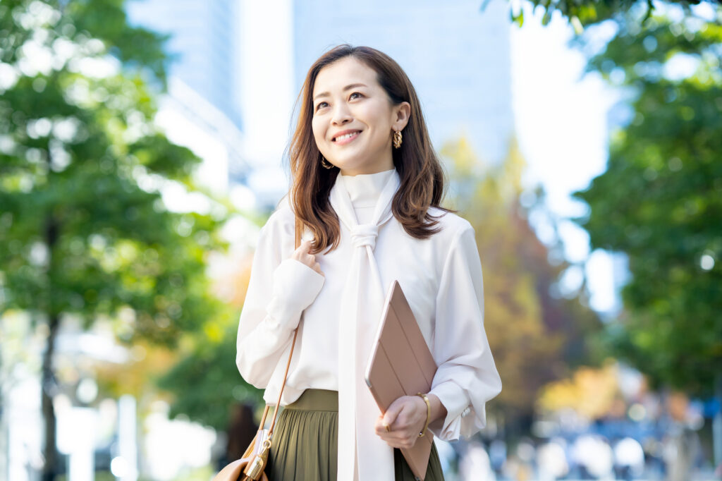 A woman in a white blouse holds a tablet on a tree-lined city street, office towers behind her