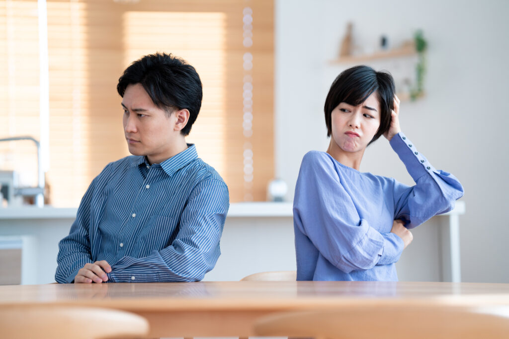 A young couple sit at a dining table looking away from each other with unhappy expressions
