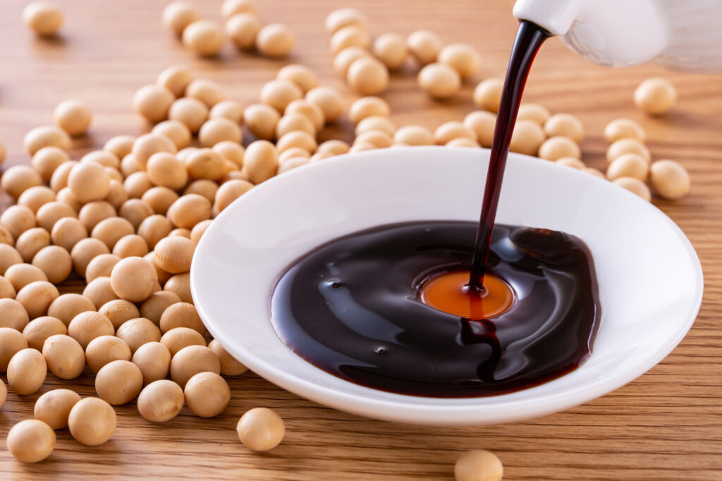 Soy sauce being poured from a white pitcher into a small dish, with dried soybeans scattered alongside.