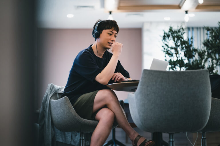 A man in a T-shirt and shorts works on a laptop, bare legs crossed, in a modern office lounge.