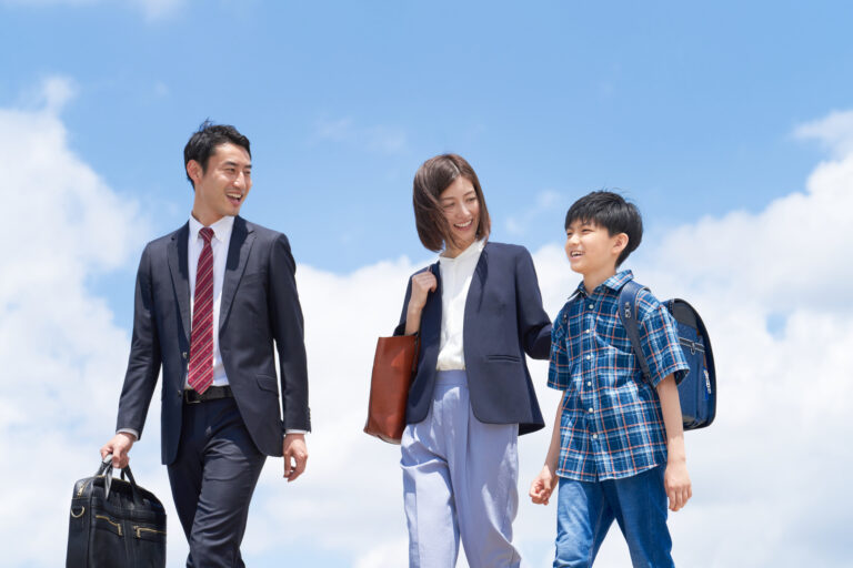 A father in a suit, a mother in a blazer, and a schoolboy with a randoseru backpack walk together under a blue sky