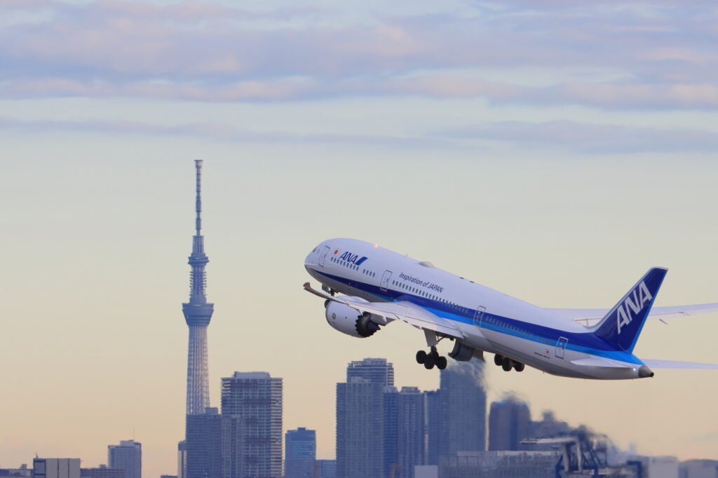 An ANA Boeing 787 climbs after takeoff past Tokyo Skytree and the city skyline at dusk