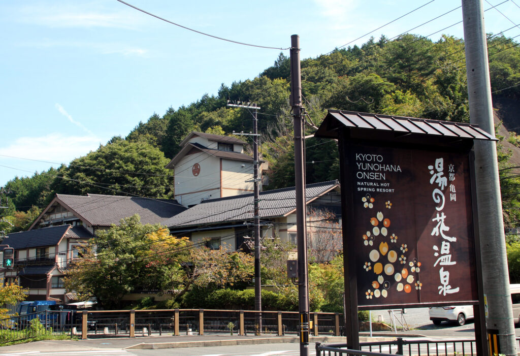 Roadside sign for Kyoto Yunohana Onsen hot spring resort, with traditional ryokan buildings and forested hills behind