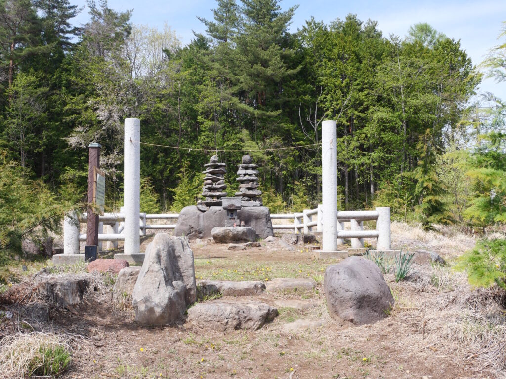 Two stacked-stone pagodas atop a low boulder at Shirahata Shrine, ringed by rope and white posts in a forest clearing