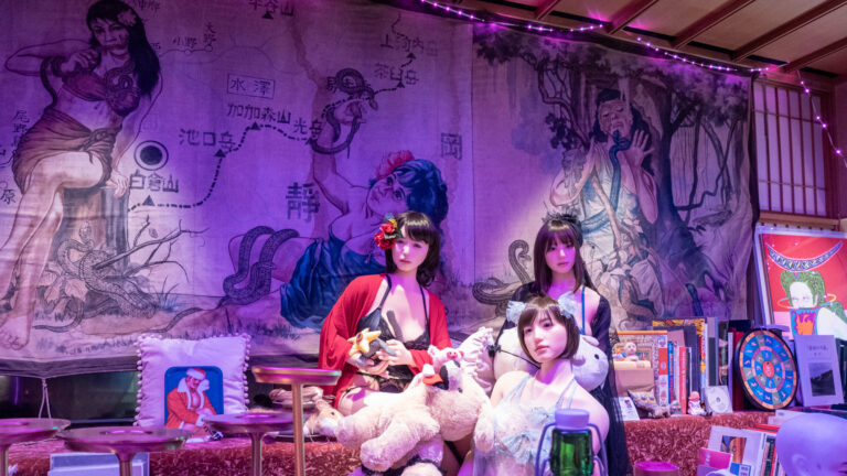 Three lifelike love dolls in lingerie seated among plush toys and books at a bar, before a purple-lit painted mural