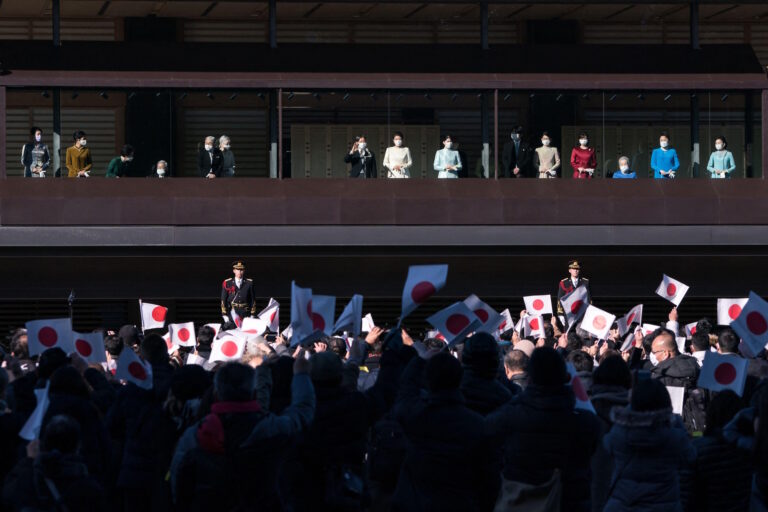 Japan's imperial family waves from a palace balcony to a crowd holding Japanese flags below.
