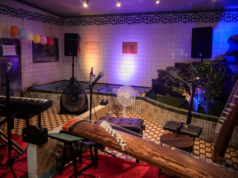 A koto, keyboard and microphones set up in a former bathhouse's tiled bathing room, paper lanterns strung above the empty tubs.
