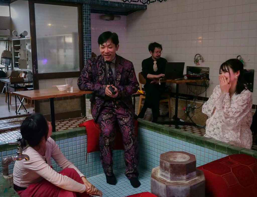 A performer in a patterned suit speaks to audience members seated around an empty tiled bathtub at Kujo-yu.