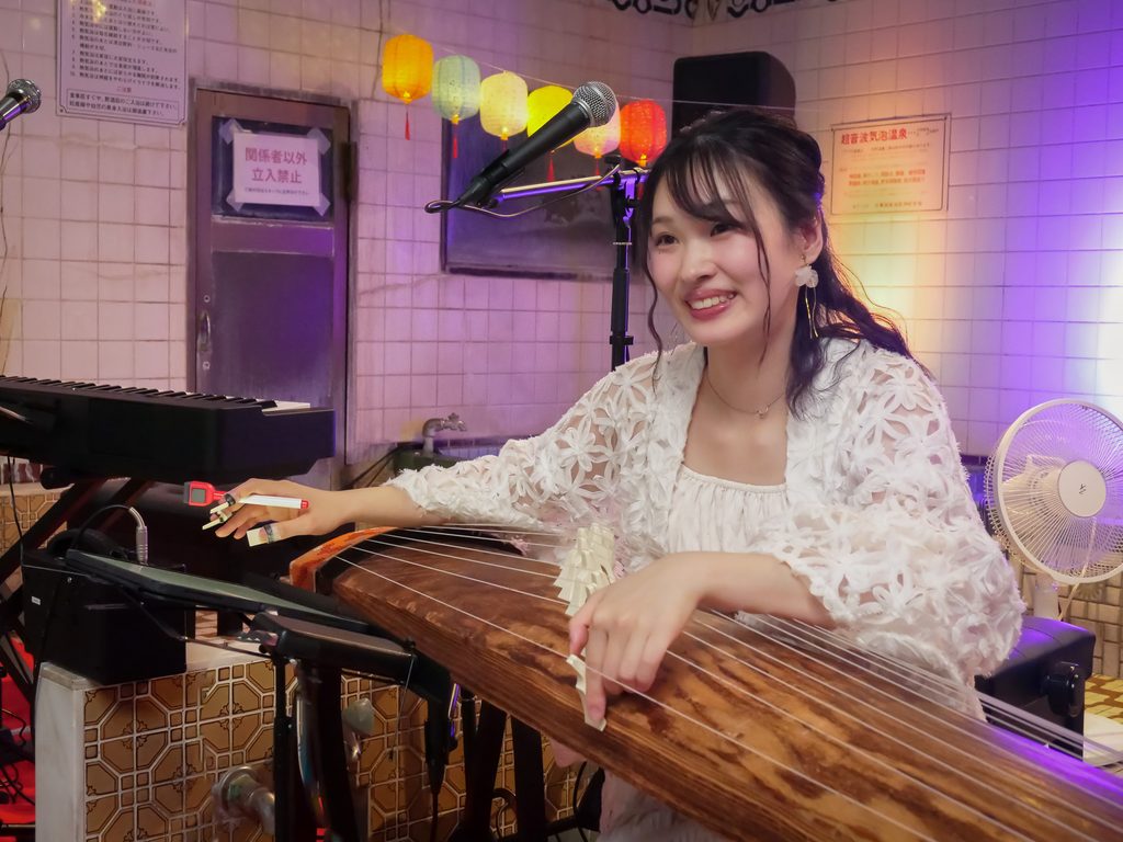 A musician wearing picks on her fingers plays the koto beside a keyboard, under paper lanterns in the tiled bathhouse.