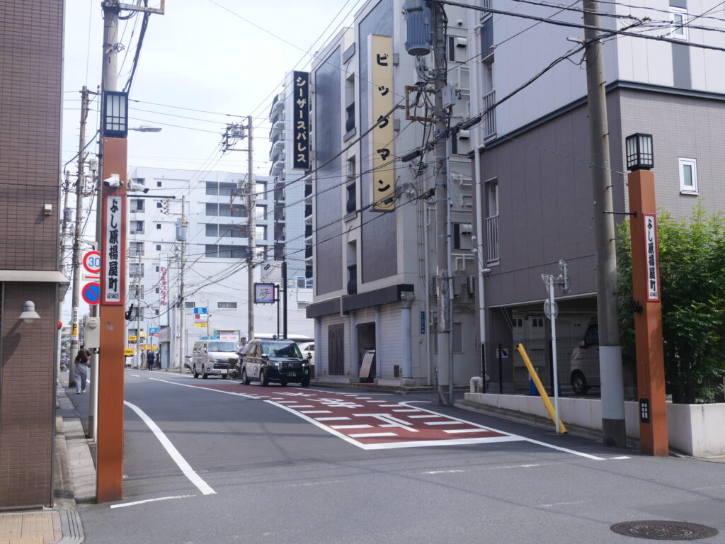 A Yoshiwara street corner marked by two wooden pillars reading “よし原” (Yoshiwara), with plain, unmarked buildings nearby.