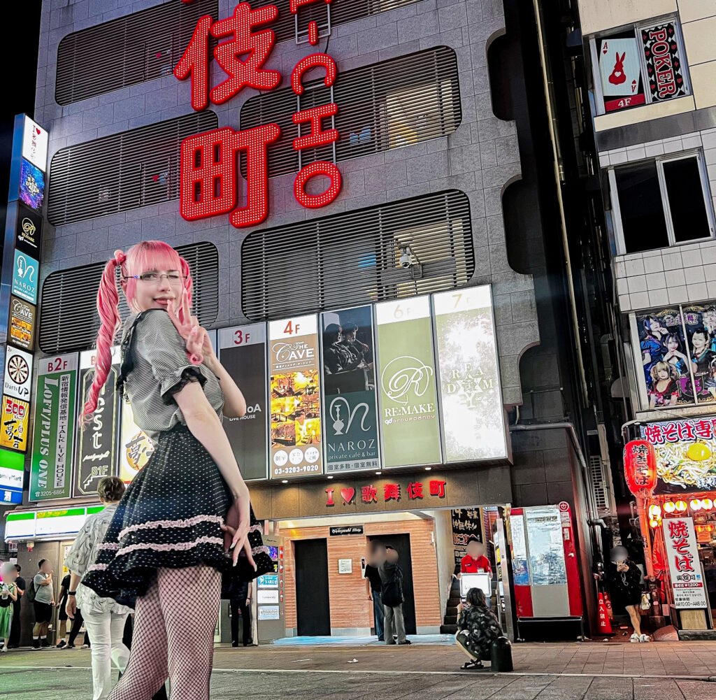 A person with pink twin-tails and a frilly black dress posing beneath the illuminated Kabukicho sign in Shinjuku at night